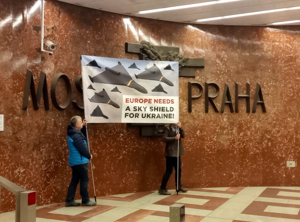 Activists covering sculpture Moscow-Prague on Prague metro station Anděl by large banner with drone swarms and text EUROPE NEEDS A SKY SHIELD FOR UKRAINE! - this second picture is showing that activists were moving between Moscow-Prague large words of the sculpture - imitating drones flying in this direction, it was acompanied by Shahed drone sound called "moped" by people in Ukraine.