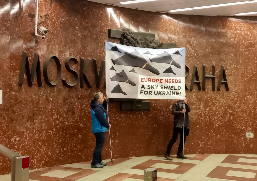 Activists covering sculpture Moscow-Prague on Prague metro station Anděl by large banner with drone swarms and text EUROPE NEEDS A SKY SHIELD FOR UKRAINE! - this third picture is showing that activists were moving between Moscow-Prague and final destination was covering part of "Prague" word of the sculpture. Then they move back and again until the station guard stopped the show and send activist away.